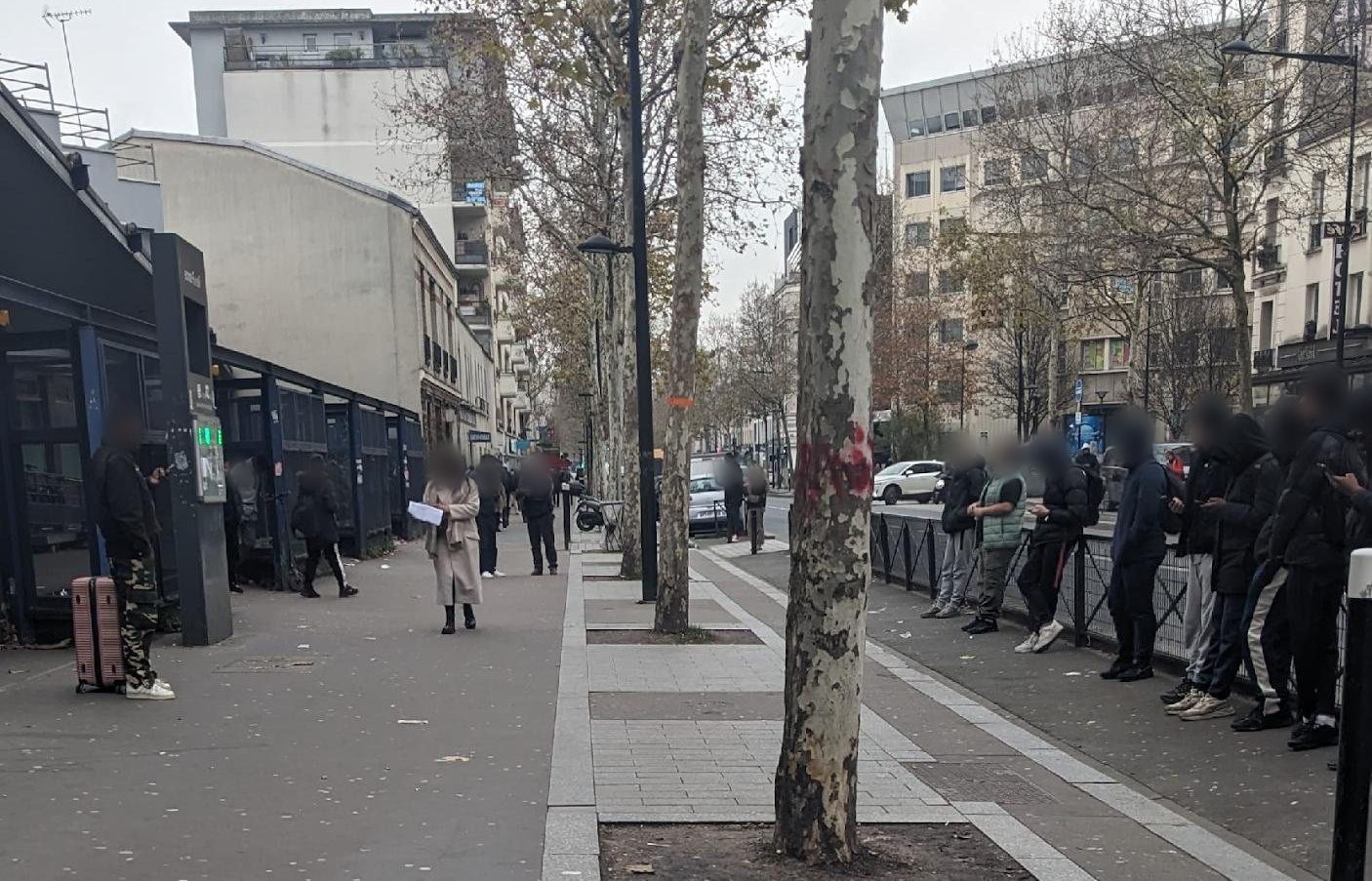 Trottoir de l'avenue de la République à Aubervilliers, près d'une sortie de métro : alignement d'arbres, piétons et groupe de jeunes le long d'une barrière